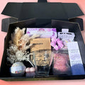 Gift box with decorative items including a glass mug, flowers, and a card on a pink background.