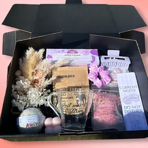 Gift box with decorative items including a glass mug, flowers, and a card on a pink background.